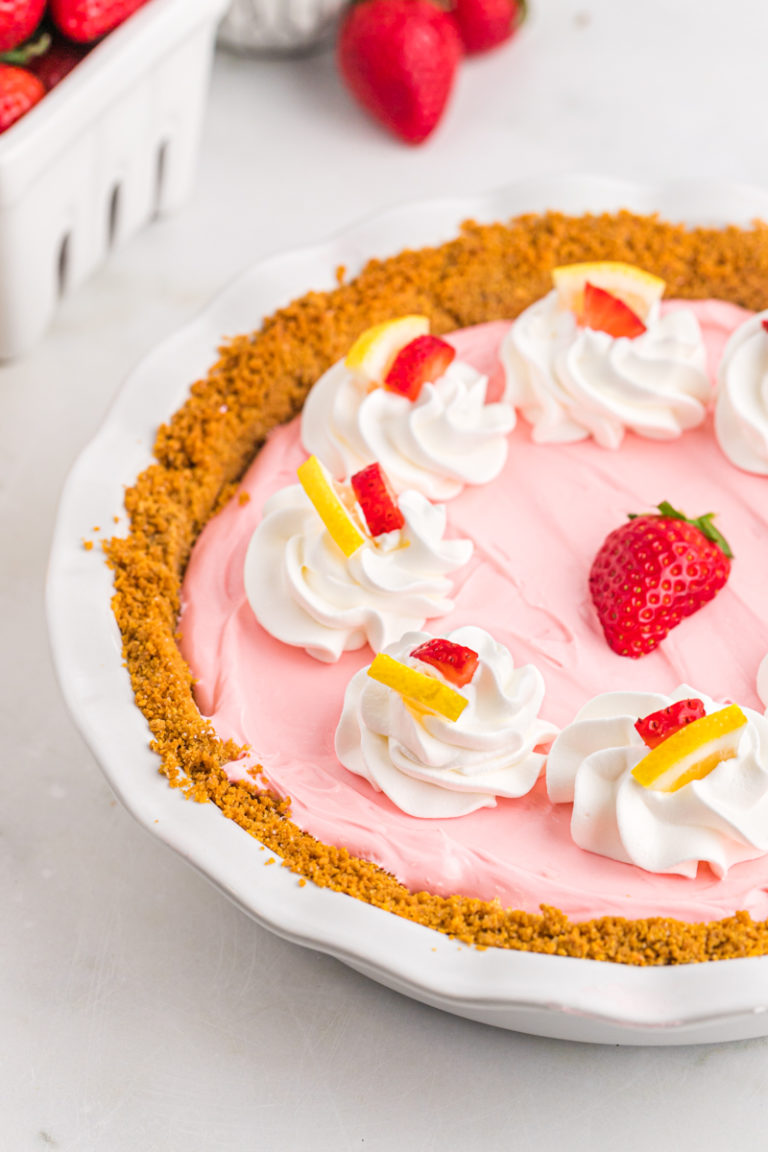 A deliciously pink no bake Kool-Aid pie with garnishes of whipped cream, lemon wedges and strawberries