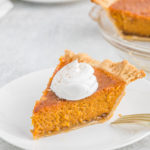 A slice of Patti Labelle's Sweet potato pie with whipped cream on a white plate with a nearby slice in the background