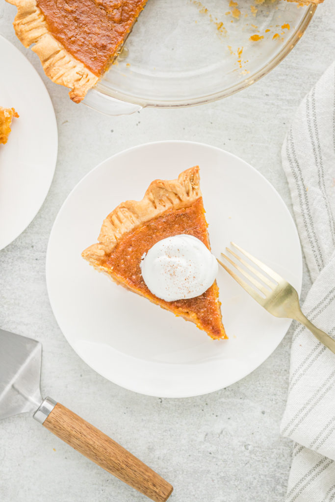 A top down shot of a sweet potato pie slice on a plate with whipped cream with the full pie nearby.