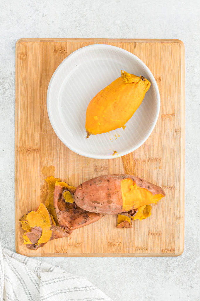 One peeled sweet potato and one nearby being peeled on a wooden board