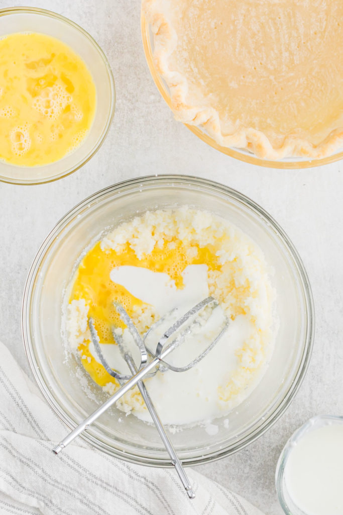 Butter and sugar being created together in a clear bowl next to a pie crust before baking.