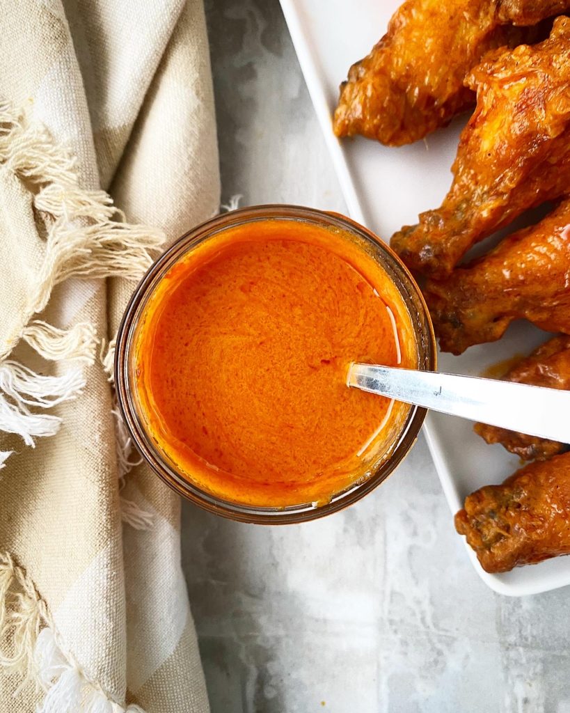 Overhead shot of Homemade Buffalo Sauce in a jar with Buffalo wings on a white plate to the right side