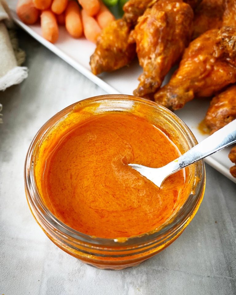 Homemade Buffalo Sauce in a jar & spoon. Buffalo wings on a white plate in back