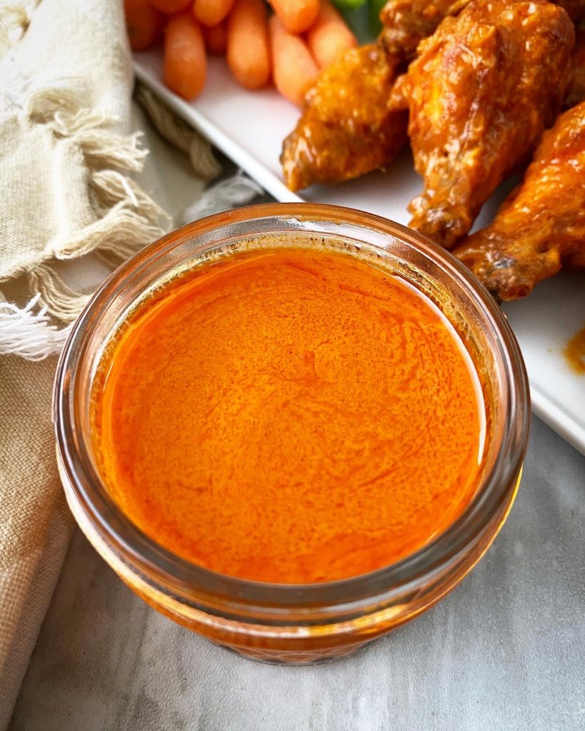 Homemade Buffalo Sauce in a jar with Buffalo wings on a white plate in back