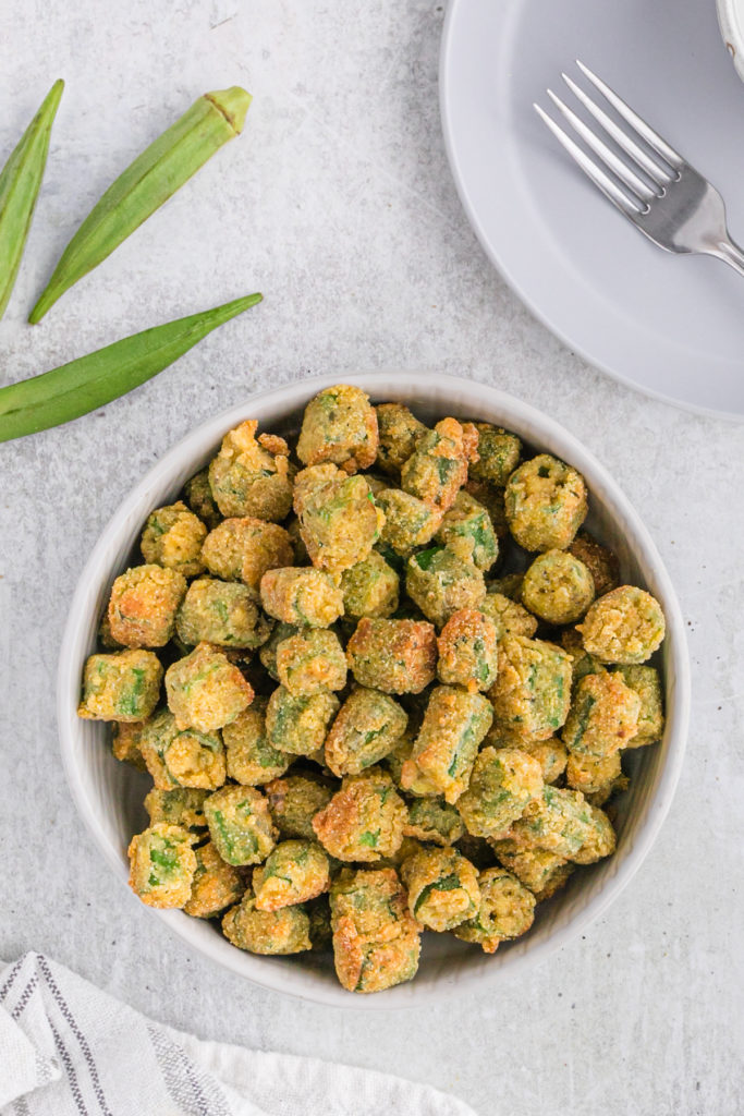 A white bowl of Southern fried okra with fresh okra nearby.