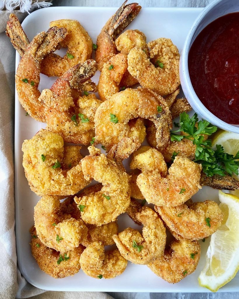 close up of fried shrimp on a white plate with lemon wedges & cocktail sauce in a bowl