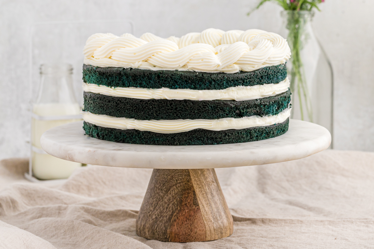 A layer cake with cream cheese frosting and blue layers on a white and wood cake stand
