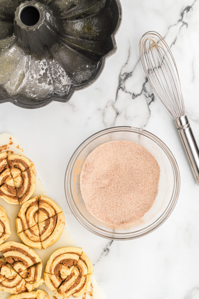 A clear bowl of cinnamon sugar next to cuts of cinnamon roll dough