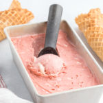 A red velvet ice cream being scooped from a frozen canister with waffle cones surrounding it