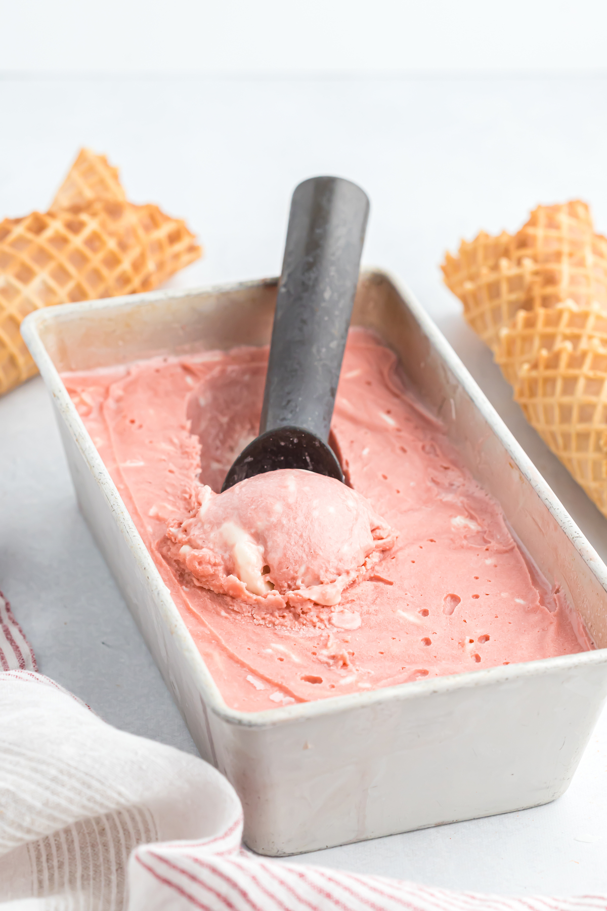 A red velvet ice cream being scooped from a frozen canister with waffle cones surrounding it