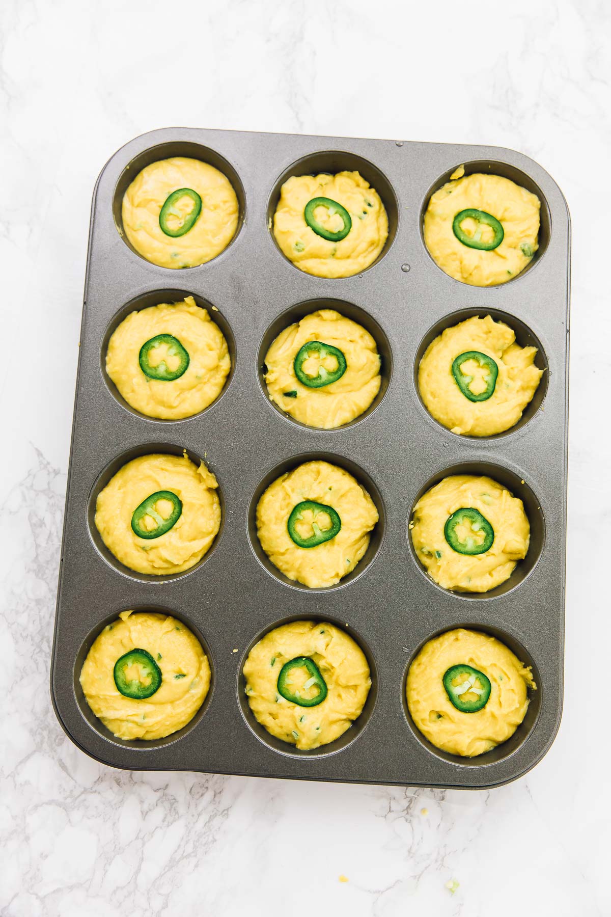 A muffin tin filled with unbaked vegan cornbread muffins, with jalapeño slices on top