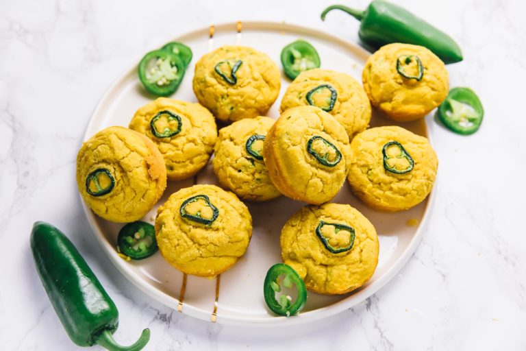 A plate full of vegan cornbread muffins, with jalapeño slices on top, and raw jalapeño slices on the plate