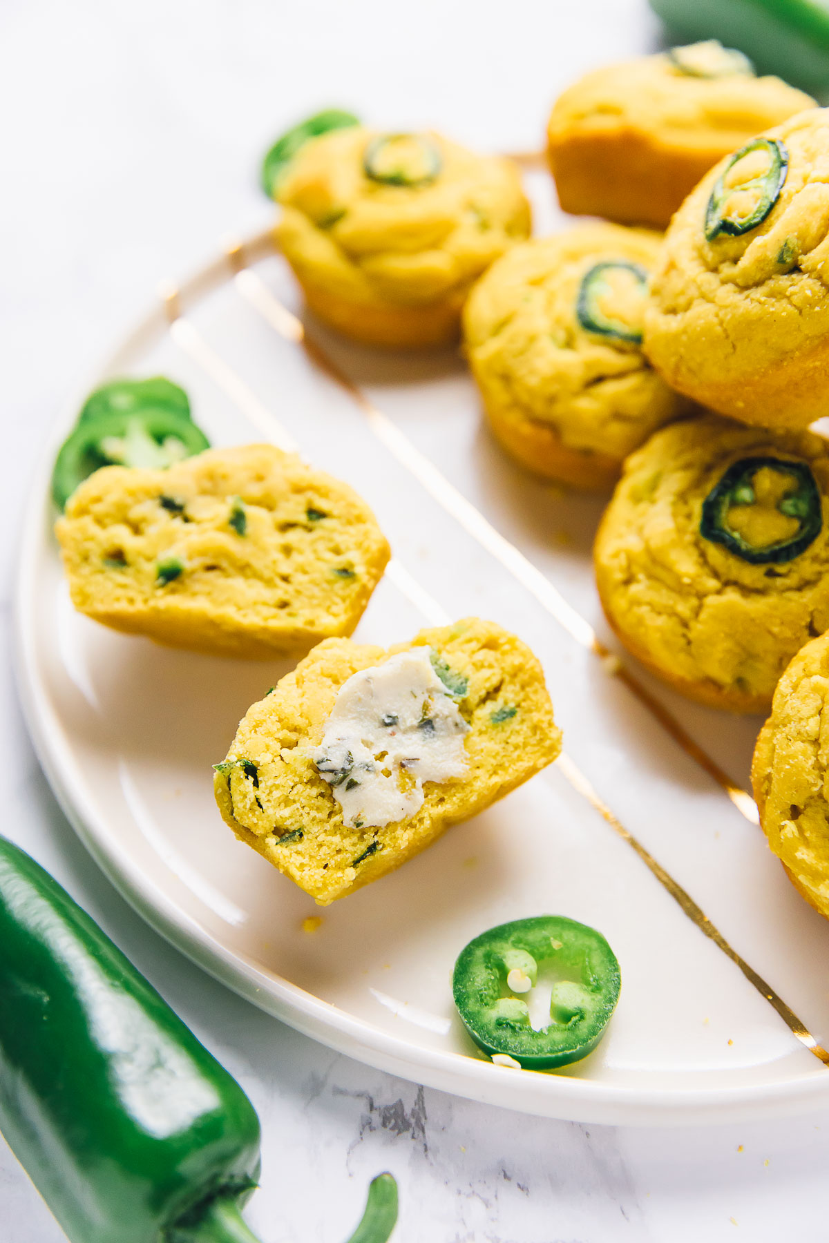 Close up of a plate of vegan cornbread muffins, with one cut in half with vegan butter on it, with sliced jalapeños surrounding it