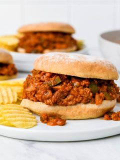 A vegan sloppy joe sandwich on a white plate with potato chips, and more sloppy joe sandwiches in the background