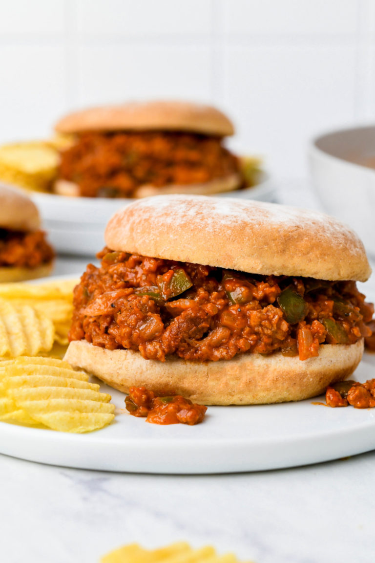 A vegan sloppy joe sandwich on a white plate with potato chips, and more sloppy joe sandwiches in the background