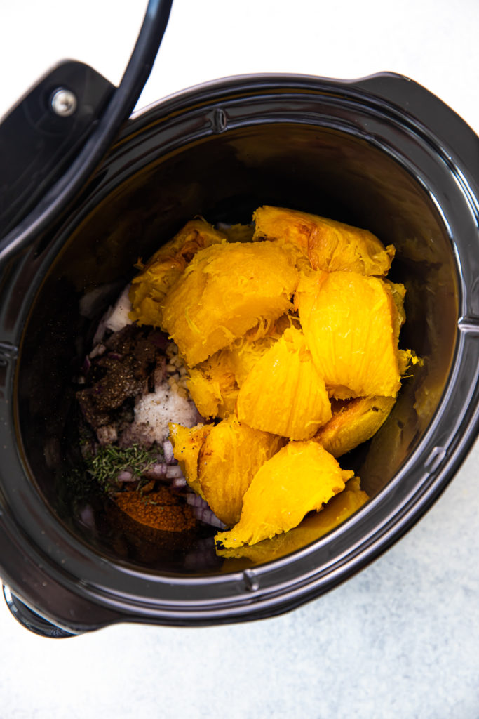 A slow cooker with uncooked pumpkin, thyme, garlic, onion, and spices
