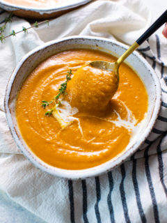 A bowl of pumpkin soup garnished with coconut milk and thyme, with a spoon taking a spoonful of soup out