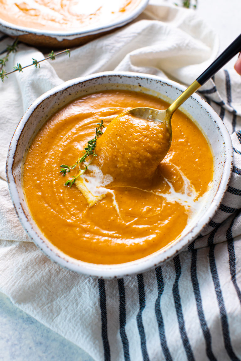 A bowl of pumpkin soup garnished with coconut milk and thyme, with a spoon taking a spoonful of soup out