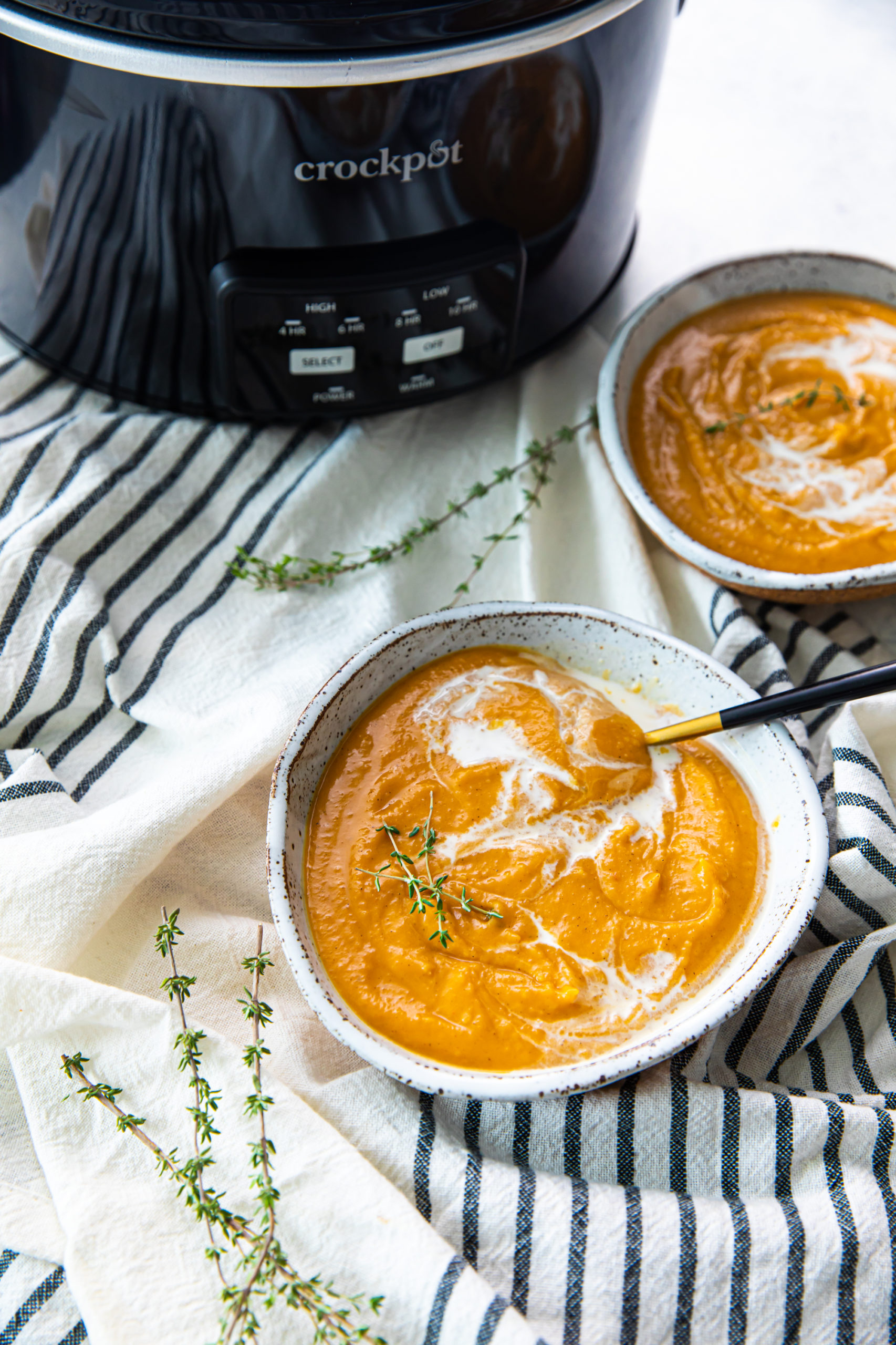 A bowl of pumpkin soup with a spoon in it, on top of a kitchen towel, next to thyme sprigs, a slow cooker, and another bowl of soup