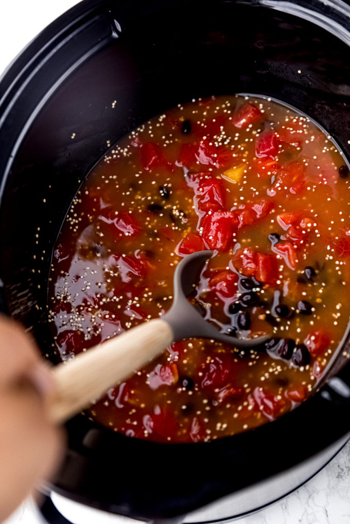 Canned tomatoes, black beans, uncooked quinoa, and vegetable broth stirred together in a slow cooker, with a stirring spoon in the slow cooker