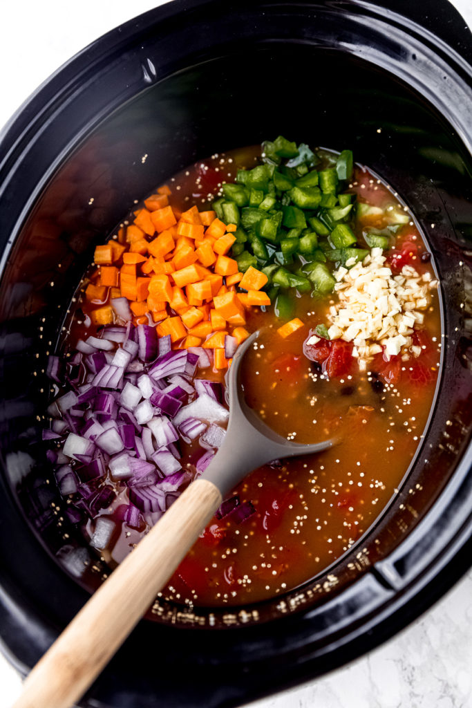 Red onions, carrots, greenbell peppers, and garlic on top of a vegan chili base in a slow cooker, with a stirring spoon