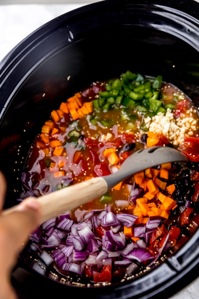 A stirring spoon stirring carrots, red onions, green bell peppers, and garlic into chili in a slow cooker
