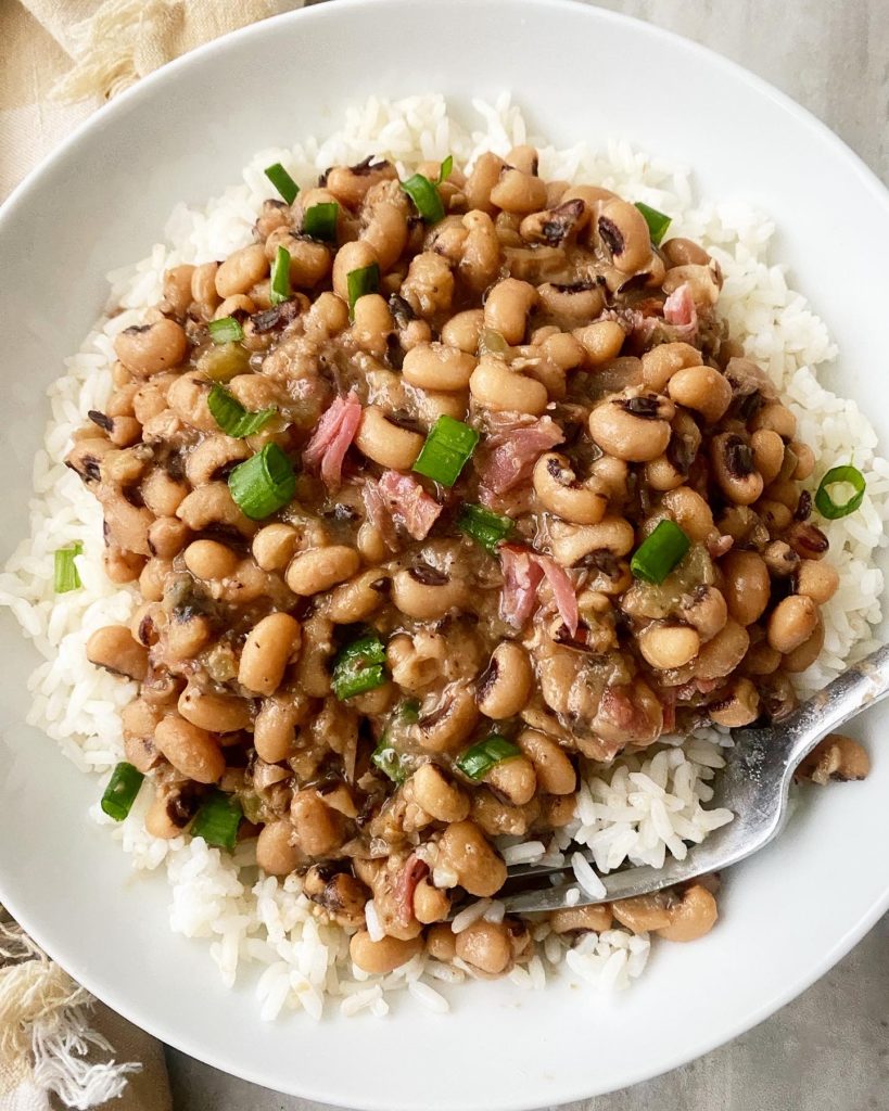 overhead shot of cooked hoppin john over rice in a white bowl garnished with sliced green onions with a silver spoon