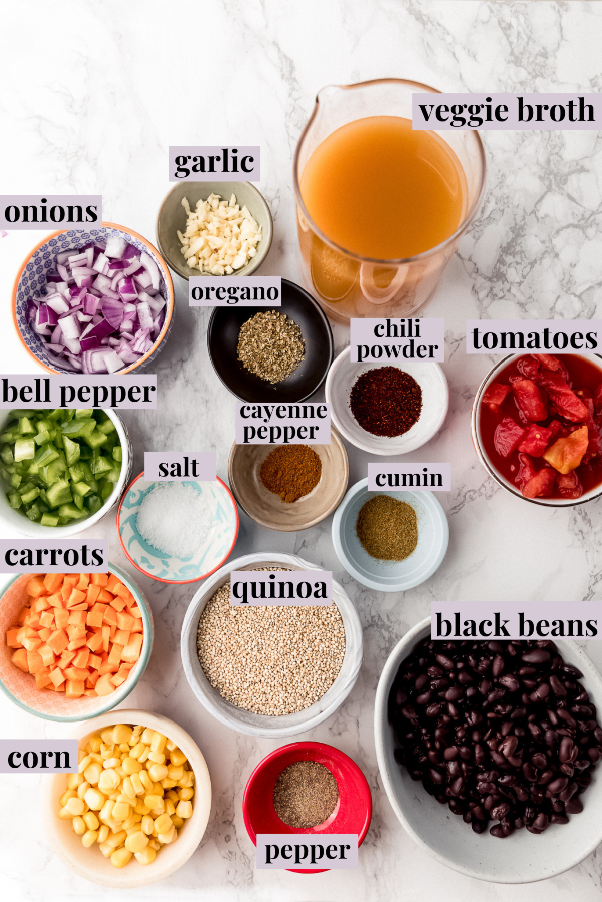 Overhead view of the ingredients for vegan chili, in bowls, labeled: veggie broth, garlic, onions, bell pepper, salt, tomatoes, chili powder, cayenne pepper, oregano, cumin, carrots, quinoa, corn, pepper, black beans