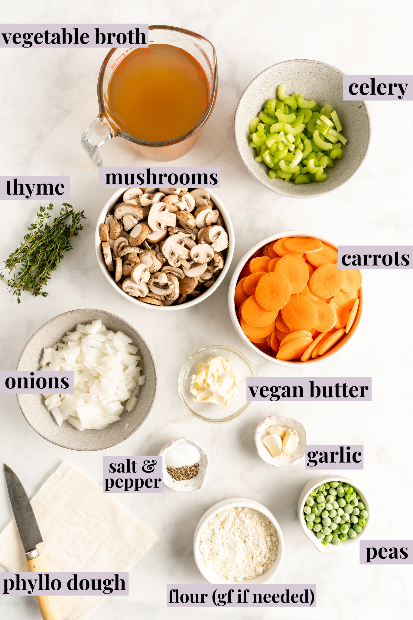 Overhead view of the labeled ingredients for vegan pot pie: a pyrex of vegetable broth, a bowl of celery, a bowl of mushrooms, a bowl of carrots, sprigs of thyme, a bowl of onions, a bowl of salt and pepper, a bowl of garlic, a bowl of vegan butter, a bowl of peas, a bowl of flour, and a sheet of phyllo dough, with a knife on it