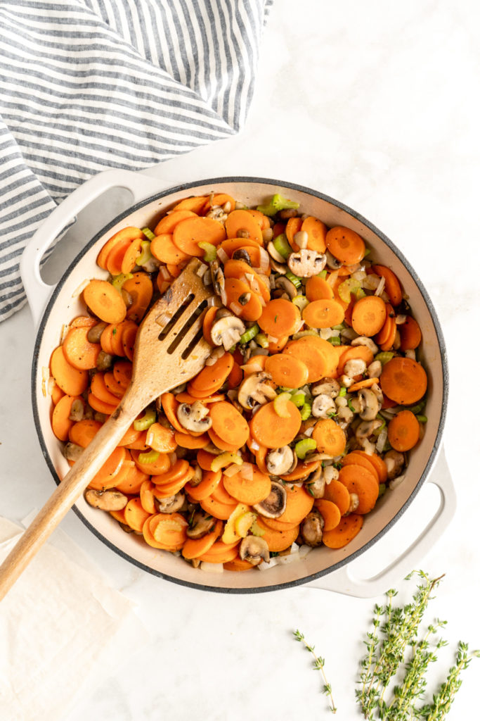 A dutch oven with carrots, onions, and celery cooking in it, with a slotted wooden spoon, next to sprigs of thyme