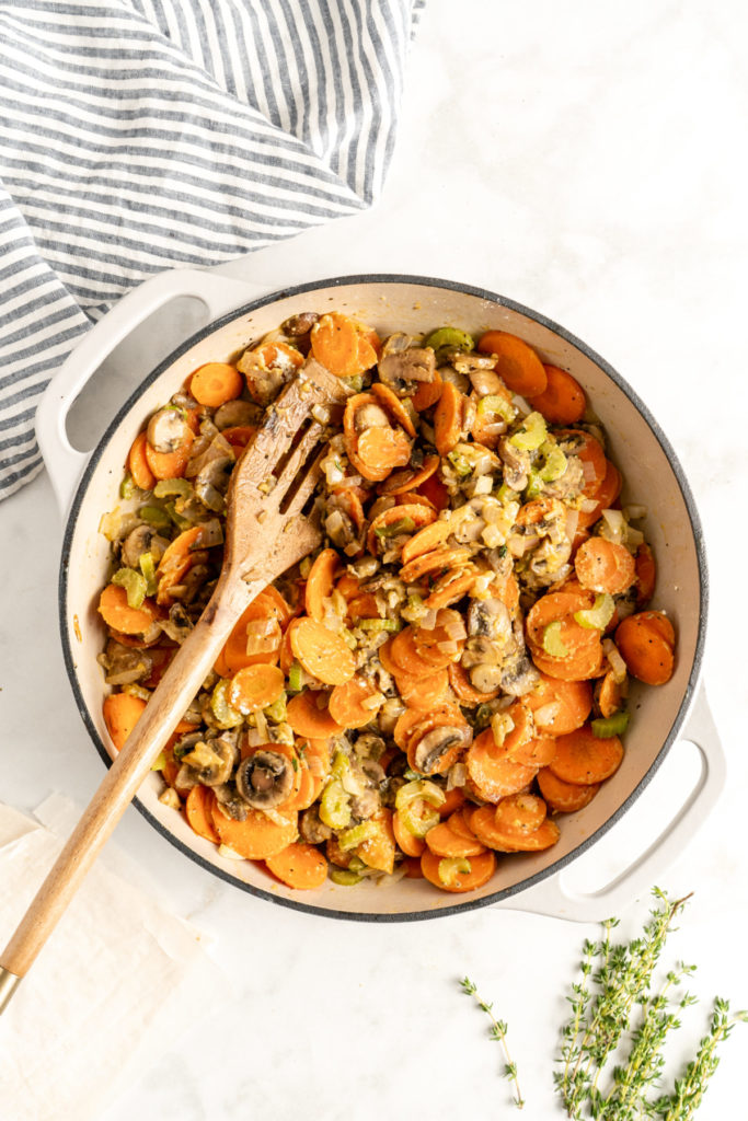 A dutch oven with carrots, celery, onions, and flour in it, with a wooden slotted spoon