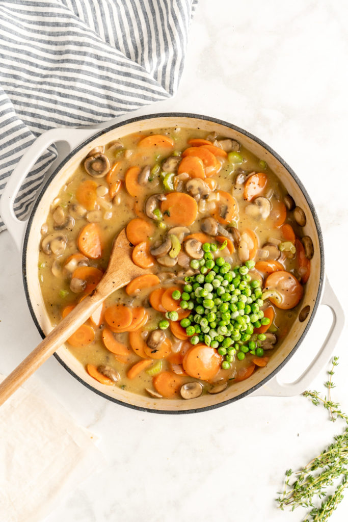 A dutch oven filled with a thick vegetable pot pie mixture, with a wooden spoon in it, and a pile of peas on top