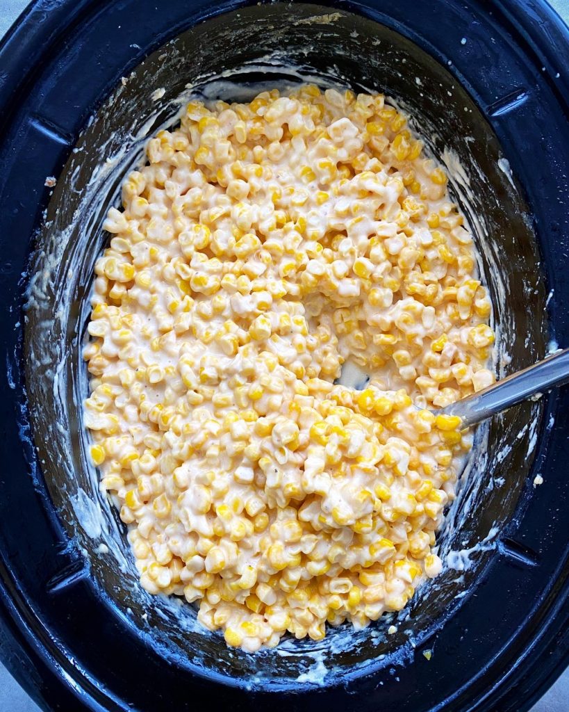 overhead shot of cooked creamed corn in a black slow cooker with a silver spoon