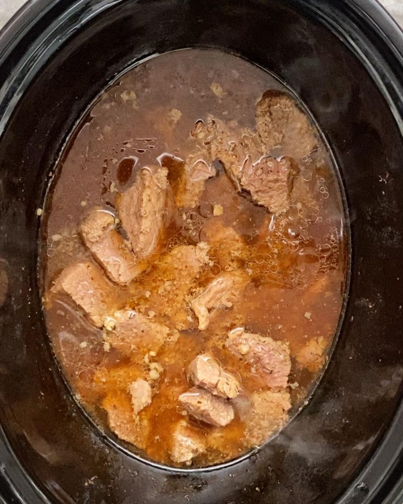 overhead shot of cooked beef tips & gravy in a black slow cooker