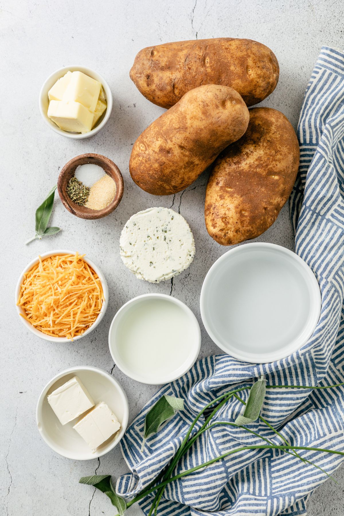 ingredients needed to make the best cheesy mashed potatoes like russet potatoes, water, salted butter, milk, garlic powder, boursin cheese, cream cheese, shredded cheese and herb seasoning in separate bowls surrounded by sage and fresh chives