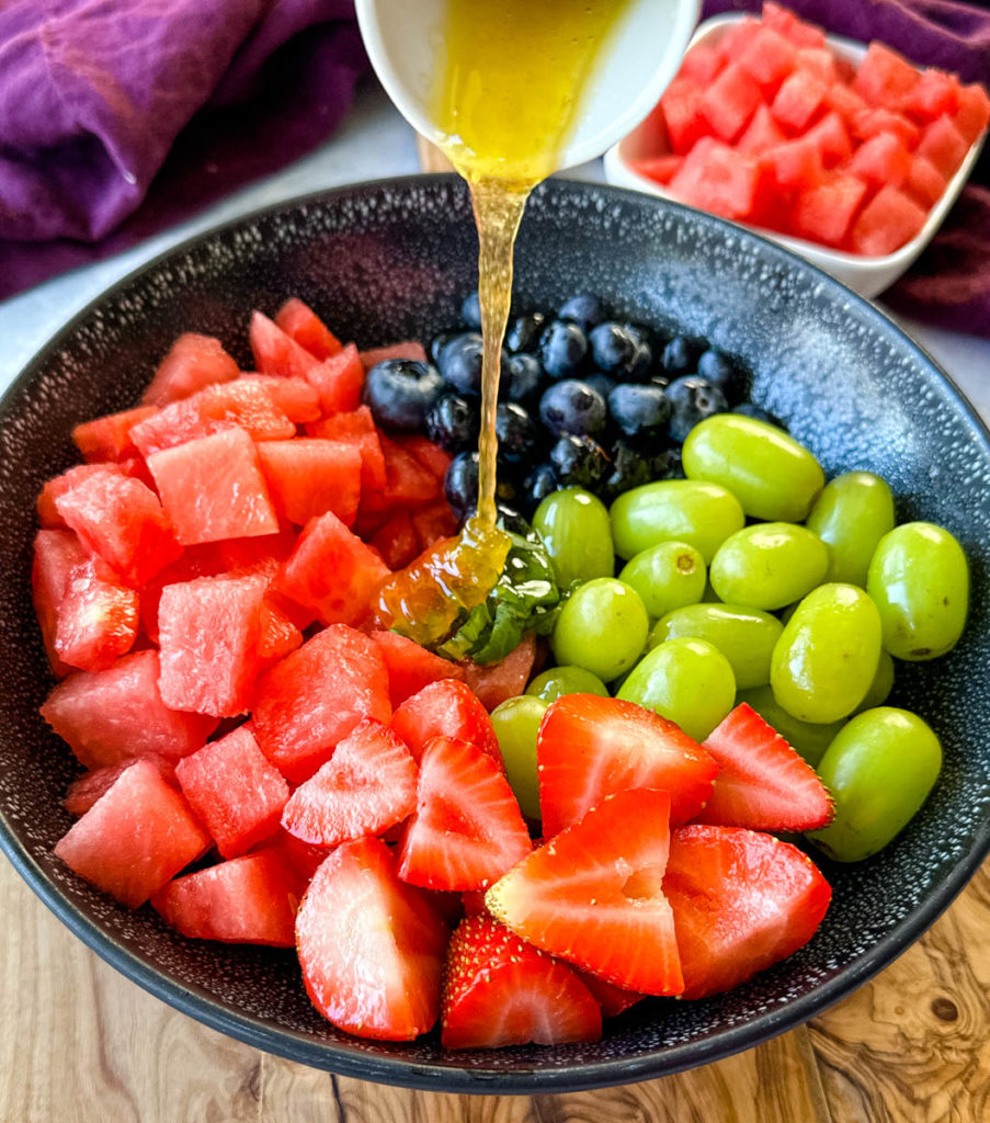 watermelon fruit salad with strawberries, blueberries, grapes, and basil in a black bowl with honey lime dressing poured over it