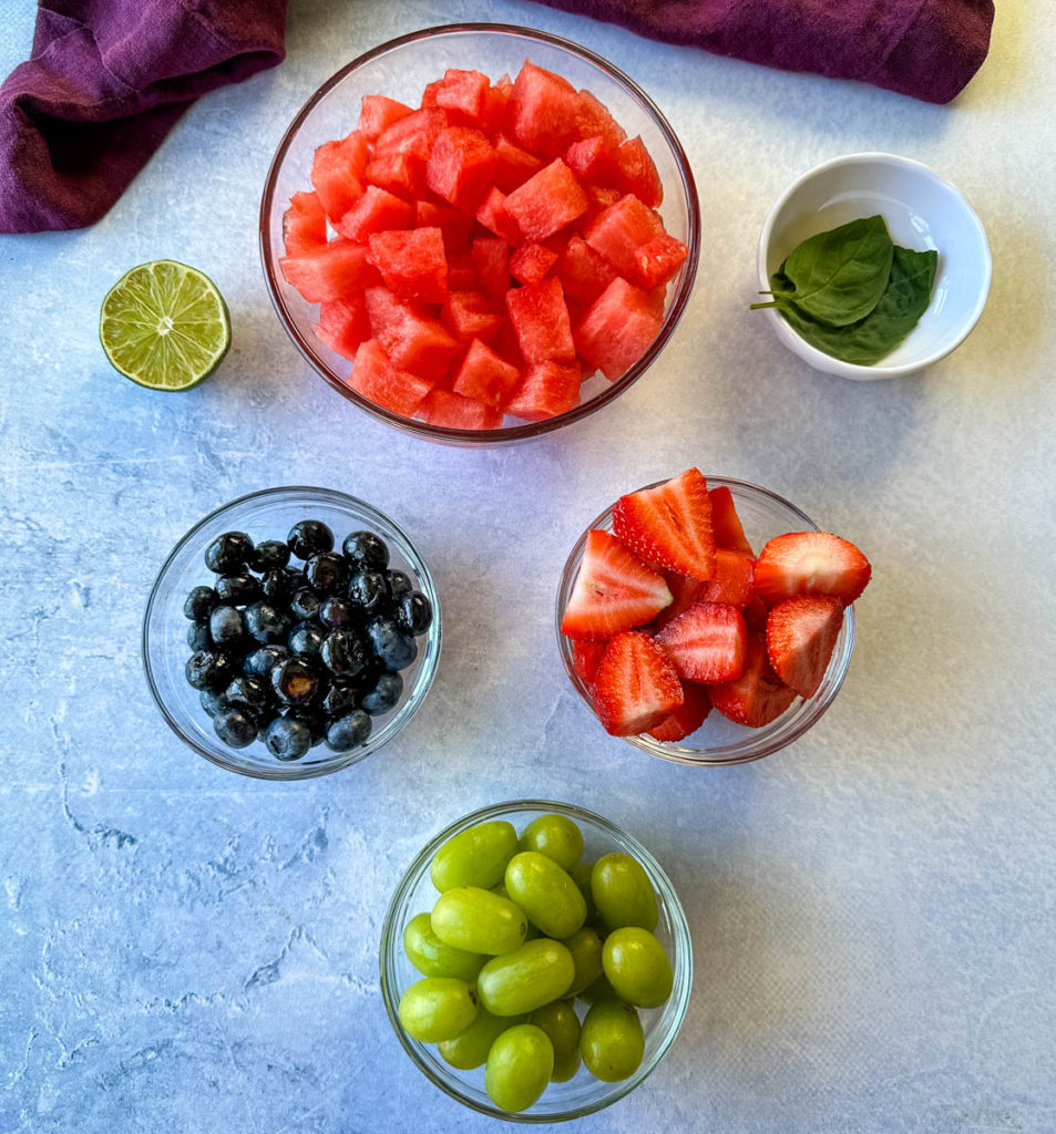 fresh watermelon, blueberries, grapes, strawberries, lime, and basil in separate glass bowls