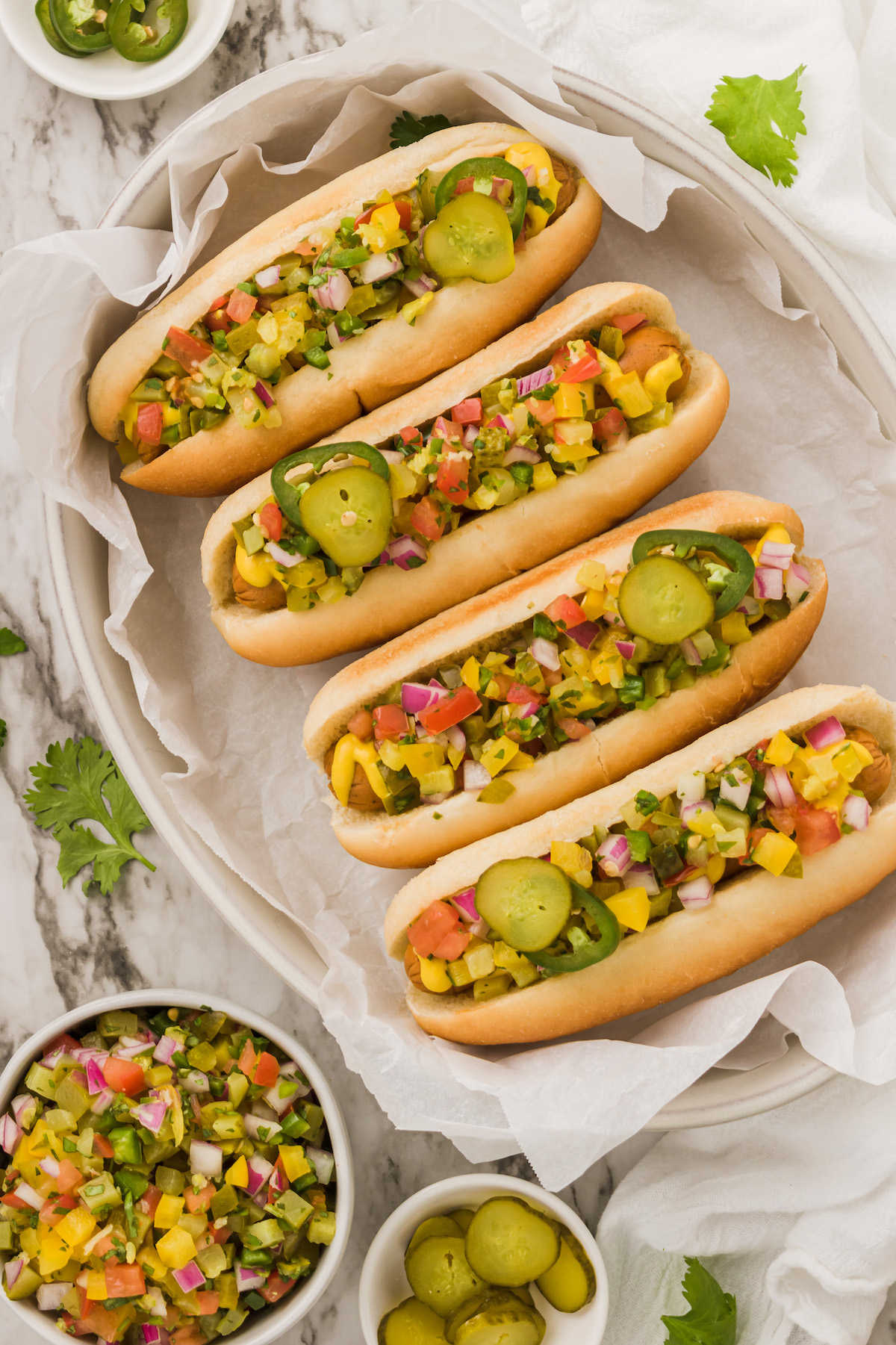 Overhead view of four hot dogs on parchment-lined platter with bowls of pickle de gallo and sliced pickles