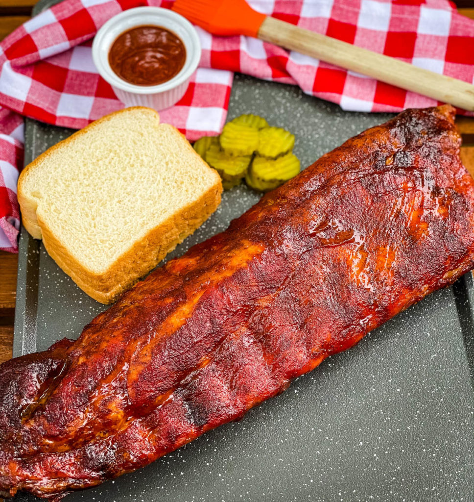 a rack of ribs on a platter with BBQ sauce, white bread, and pickles