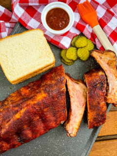 a rack of ribs sliced on a platter with BBQ sauce, white bread, and pickles