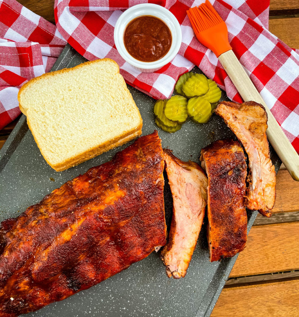 a rack of ribs sliced on a platter with BBQ sauce, white bread, and pickles
