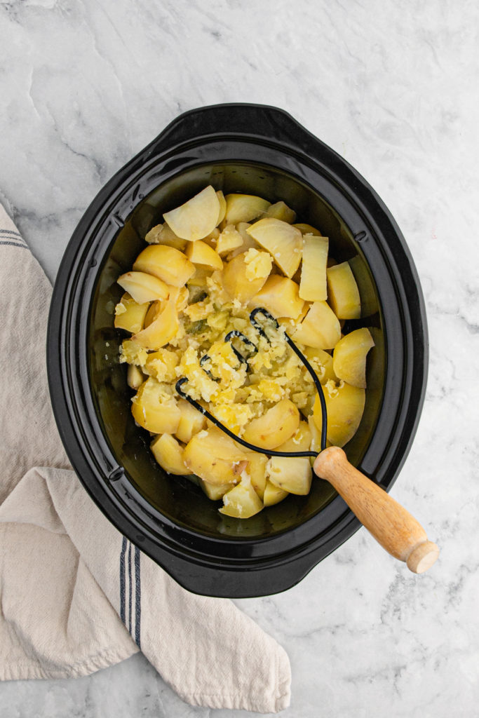 A masher mashing potatoes in a slow cooker to enjoy