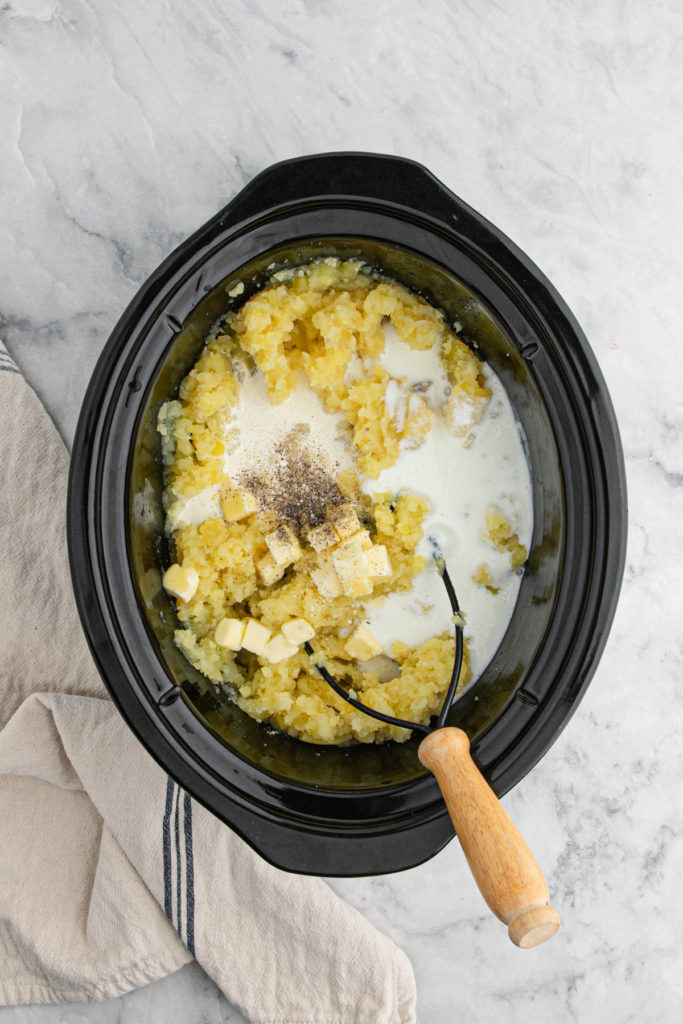 A masher mashing potatoes in a slow cooker to enjoy