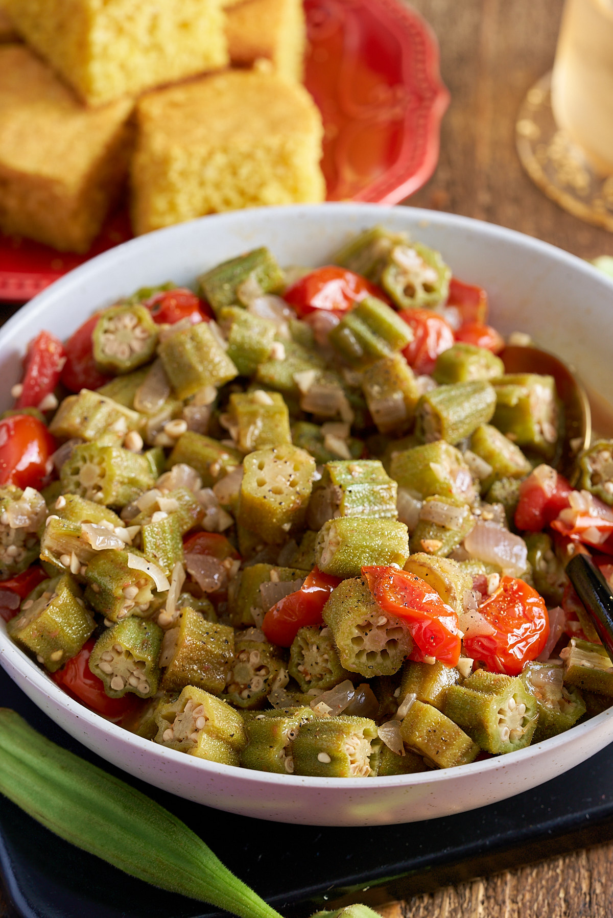 stewed okra and tomatoes in white bowl with cornbread in background on red plate