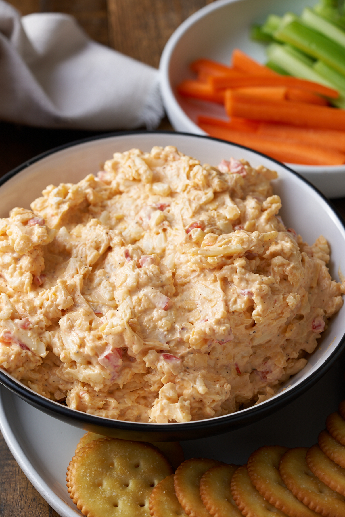 pimento cheese in white bowl with crackers on side and vegetables in background