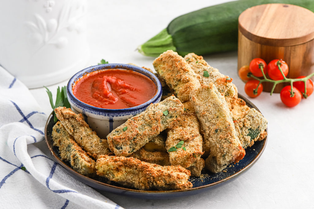 Spears of fried zucchini on a black plate with a bowl of marinara sauce