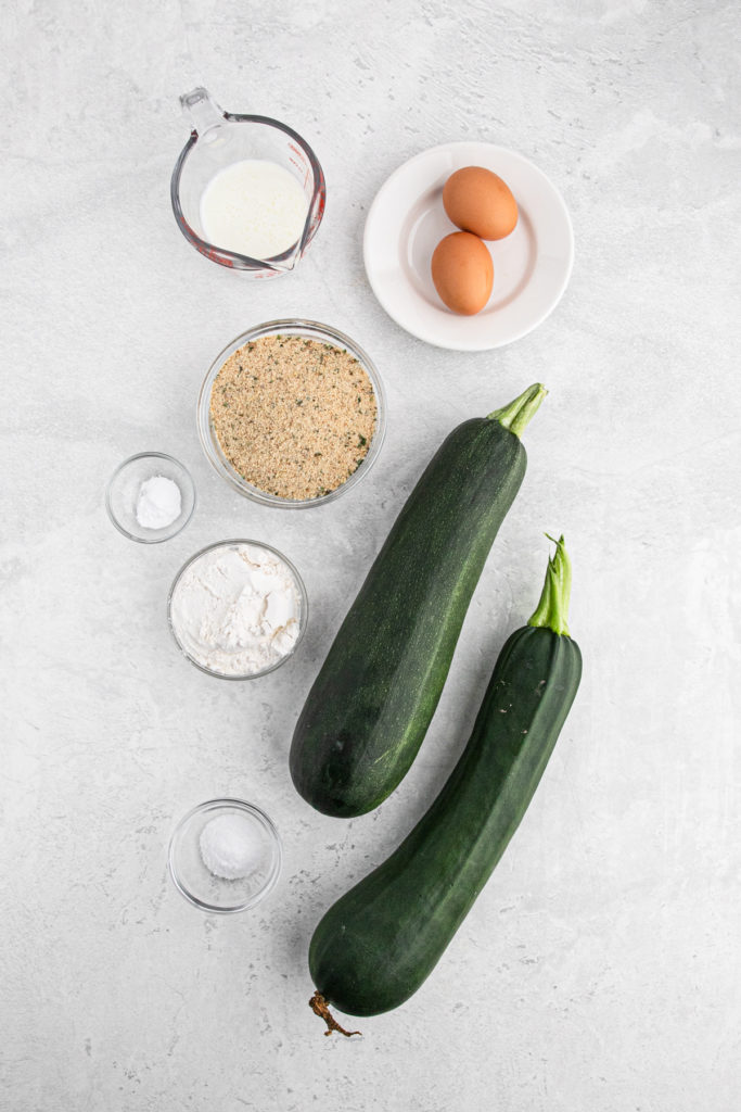 Two zucchini, bread crumbs, eggs and milk in separate bowls