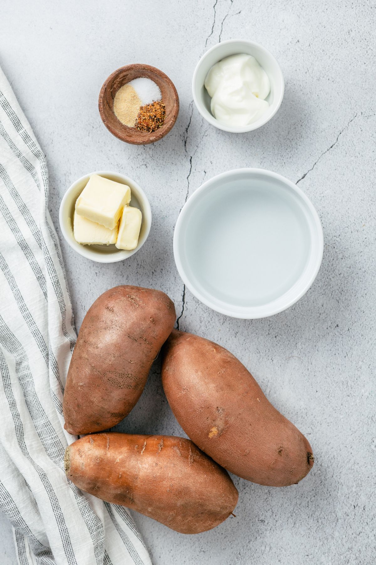 ingredients needed to make savory mashed sweet potatoes: sweet potatoes, water, butter, garlic powder, herb sprinkle seasoning, sea salt, and sour cream in separate bowls