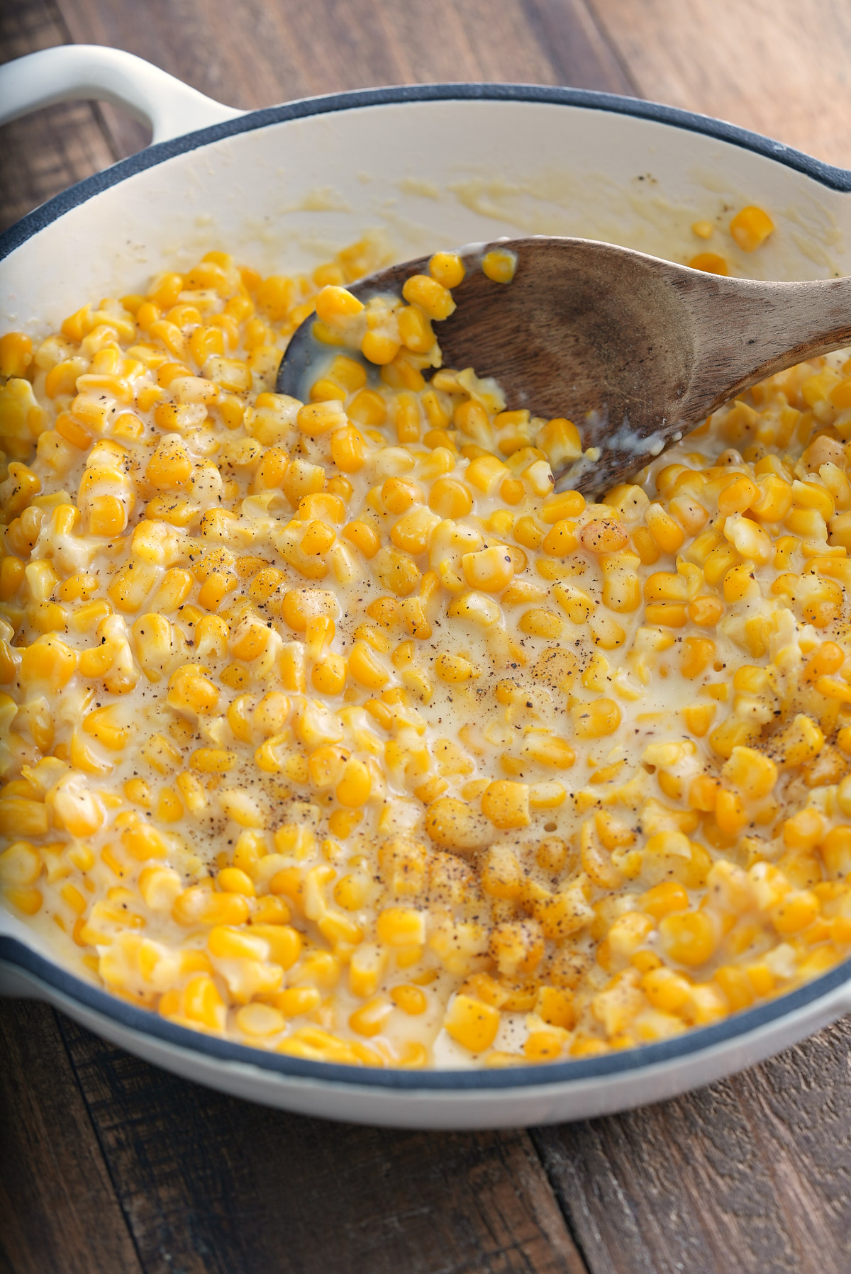 creamed corn in a white skillet with a wooden spoon scooping it up