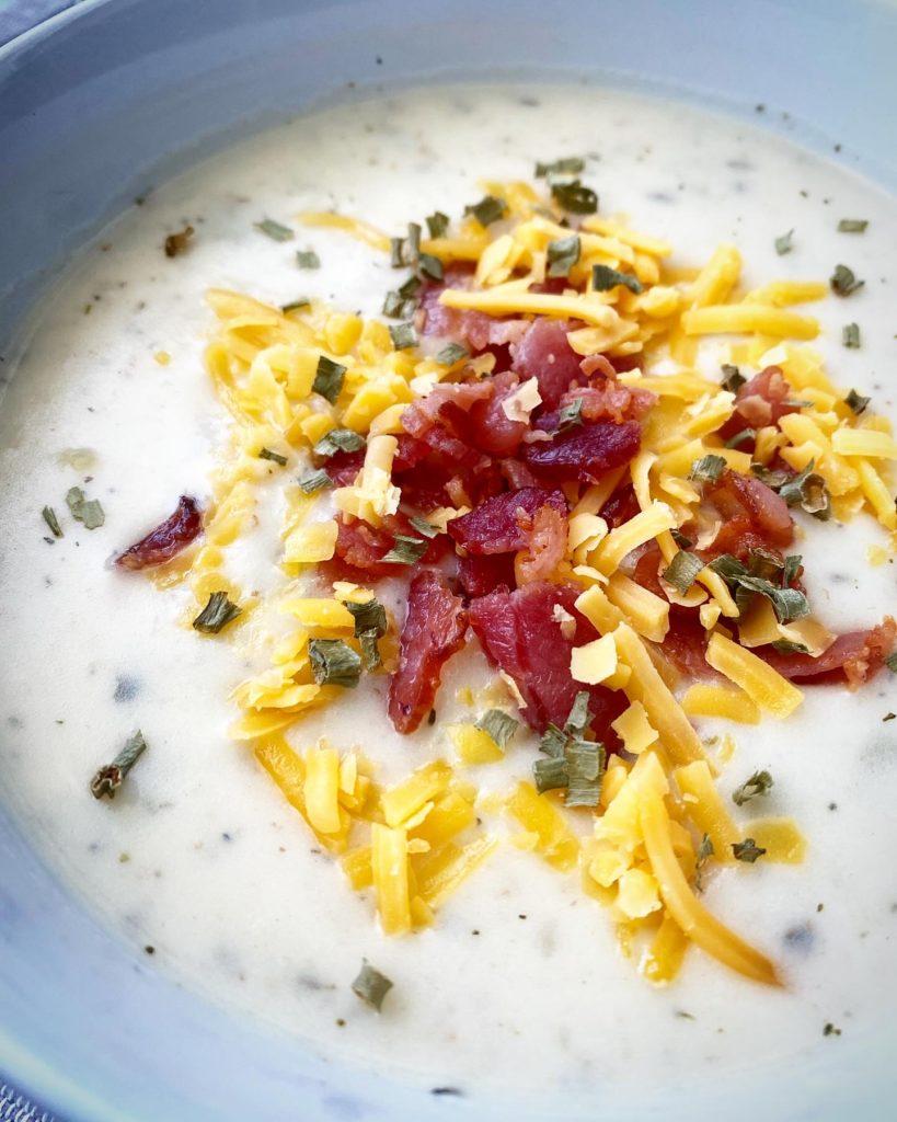 close up of potato soup in a white bowl garnished with bacon, cheddar cheese, and dried chives